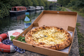 Open pizza box with a pizza inside, next to a bottle of water and a life buoy on a canal bank.